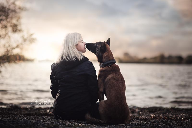 Gemeinesames Fotoshooting von Hund und Frauchen: belgischer Malinois Rüde sitzt zusammen mit einer Fraum draußen am Seeufer in Paderborn Elsen/Sande